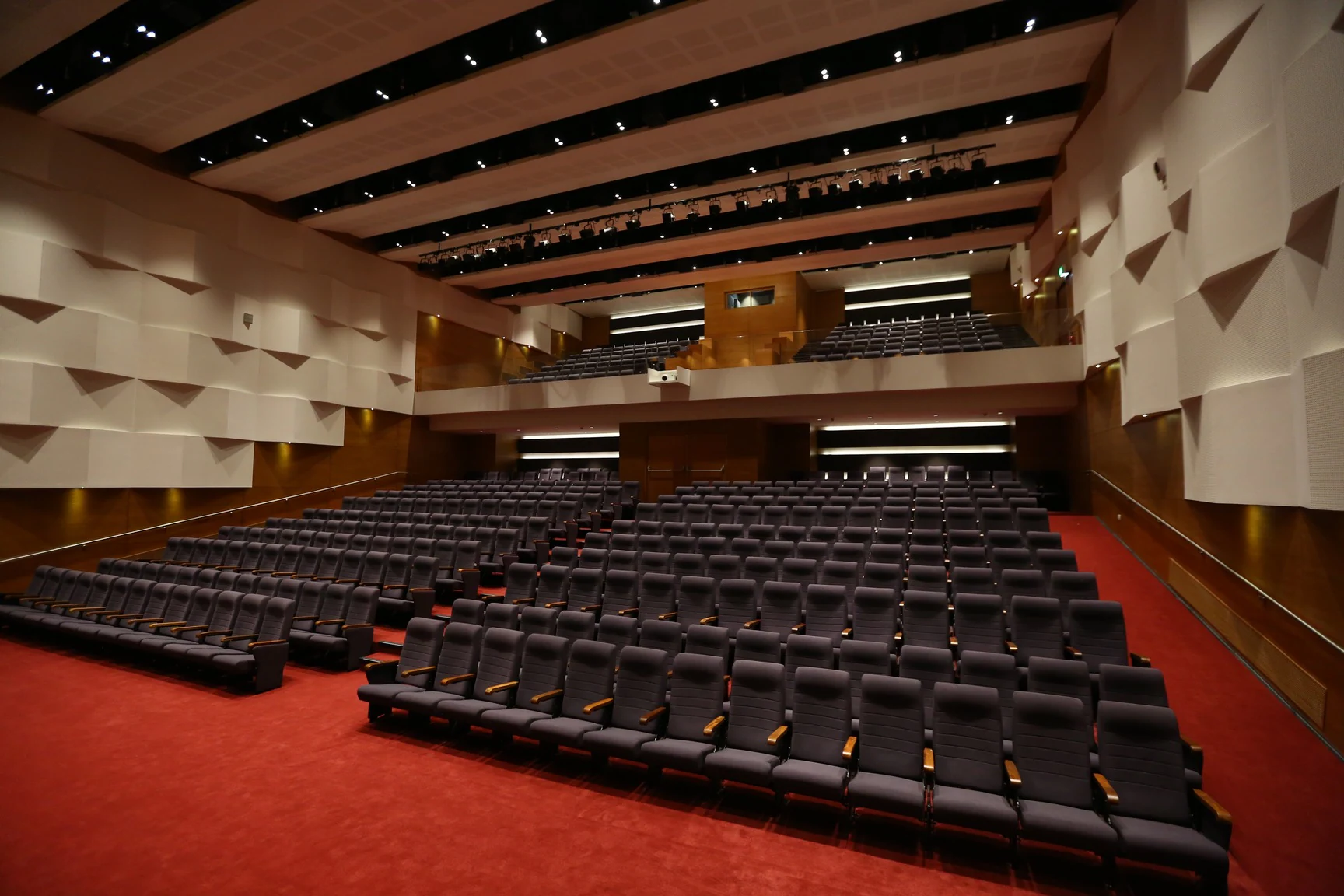 A modern, spacious auditorium with tiered seating arranged in rows on red carpeted flooring. The walls feature acoustic paneling for sound control, and the ceiling is equipped with built-in lighting and stage rigging. The auditorium includes a balcony level for additional seating and a central projector mounted on the ceiling.